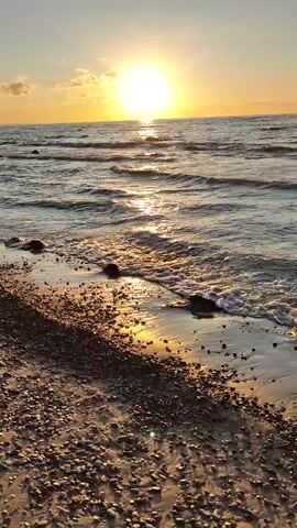 The video shows a sunset over the ocean. The sun is a bright orb near the horizon, casting a golden light on the water. Gentle waves lap at a pebble-strewn beach in the foreground. The sky is a gradient of pale blue and orange, with a few wispy clouds. The scene is peaceful and serene.