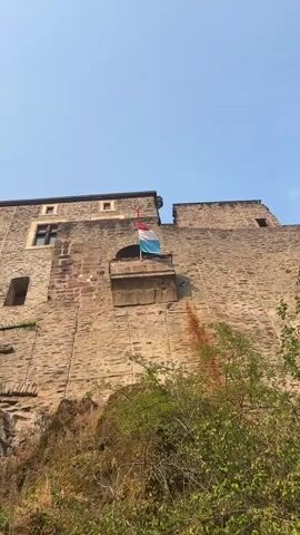 A flag with red, white, and blue horizontal stripes, likely the flag of Luxembourg, is attached to a flagpole on a small stone balcony protruding from a large, weathered stone building. The building appears to be a castle or fortress, with rough-hewn stone blocks and a few small windows visible. The flag is positioned against a clear blue sky. The camera perspective is looking up at the building from a lower vantage point, with some green foliage visible in the lower part of the frame. The building shows signs of age, with some darker staining on the stone wall below the balcony, possibly from water or rust. A portion of the building to the right of the balcony has a gap or opening. The flag is gently swaying.