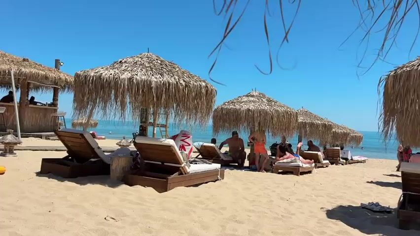 The video shows a sandy beach on a clear, sunny day with a bright blue sky. Several thatched umbrellas provide shade over lounge chairs, some of which are occupied by people. In the background, the ocean is visible with a few people in the water. To the left, there appears to be a bar area made of wood. People are seen relaxing on lounge chairs, and some are standing and interacting. One person is dancing under an umbrella. The overall scene depicts a relaxed beach atmosphere.