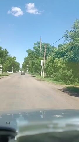 The video shows a view from inside a vehicle traveling down a paved road. The road is flanked by trees and a clear blue sky with scattered white clouds. There are utility poles with wires along the roadside. Several cars are visible on the road ahead, and one white vehicle is parked on the left side. The road appears to have some potholes. The sun is shining, indicating daylight. The video appears to be a recording of a drive through a suburban or rural area.