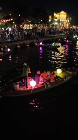 The video shows a night scene on a river filled with boats carrying passengers and illuminated by lanterns. The boats are traditional in style, with the one in the foreground being rowed by a person wearing a green shirt and a white hat. The boat has three passengers, all wearing orange life vests, and a large round lantern. The river reflects the lights of the lanterns and the buildings on the shore, creating a colorful spectacle. On the shore, a crowd of people is visible, watching the boats. The buildings along the shore are lit up with warm lights, and some are decorated with lanterns. The overall atmosphere is festive and tranquil.