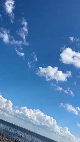 The video shows a tilting shot of a bright blue sky with scattered white clouds. In the bottom left corner, a sliver of dark blue water is visible, suggesting a coastal or oceanic environment. The clouds are varied in shape and density, with some appearing wispy and others more substantial. Sunlight glints off some of the clouds, indicating a sunny day. A faint contrail from an aircraft is visible across the upper right portion of the sky. The camera movement is a slow, upward tilt, revealing more of the sky as the video progresses.
