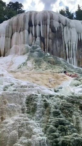 A person in swimwear is lying on their side on a rocky, mineral-covered slope where water is flowing down, creating a cascading effect. The person is positioned in the upper right portion of the frame. The slope is textured with mineral deposits, giving it a layered and somewhat icicle-like appearance in places, with shades of white, green, and yellow. The water creates small waterfalls and pools as it descends. Trees are visible in the upper background.