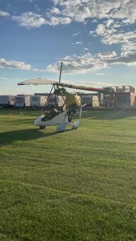 The video shows an ultralight aircraft on a grassy field. The aircraft is white with a red and yellow stripe on the wing. Two people are seated in the aircraft. The aircraft is facing to the left. In the background, there are several RVs and trailers parked in a line. The sky is blue with some white clouds and the sun is shining. The overall scene appears to be a campground or an airfield.