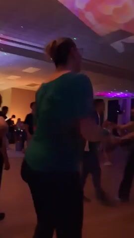 This video captures a dimly lit event space with multiple people dancing. Colorful lights, including disco balls and projected patterns on the ceiling, create a vibrant atmosphere. In the foreground, a person in a teal top is visible from behind, facing away from the camera. To their right, a woman in a dark top and pants is dancing. Other individuals are scattered throughout the room, engaged in dancing and socializing. The floor appears to be made of wood, and the walls are relatively dark. The overall impression is of a lively party or dance gathering.