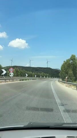 The media file captures a scenic view from a car traveling on a highway in Santa Ninfa, Italy. The road winds through a landscape dotted with lush green trees and rolling hills. In the distance, several wind turbines are visible against a bright blue sky with scattered white clouds. The sun is high, indicating it is daytime, and the weather appears clear and pleasant.

The primary subjects are the road, the surrounding natural scenery, and the man-made wind turbines. There are no visible people in the frame, but the perspective suggests someone is driving or is a passenger in the vehicle. The road itself is paved and has white lane markings. Guardrails line the sides of the highway, and prominent chevron signs indicate curves in the road.

The activity is transportation, with the camera providing a first-person view of the journey. The scene evokes a sense of travel and openness, typical of a road trip through the countryside. The location cues, such as the wind turbines and the vegetation, combined with the provided context of Santa Ninfa, Italy, paint a picture of rural Italian landscape. There is no visible text within the media file itself.