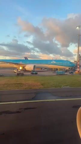 The video shows an airplane on the tarmac at an airport. The plane is blue and white with "KLM" written on its side. There are several vehicles around the plane, including what appear to be baggage carts and a tug. The sky is cloudy with some pink and orange hues, suggesting it may be sunrise or sunset. The video is filmed from inside another airplane, as evidenced by the view of an engine cowling in the lower right corner. The scene is calm and still, with no apparent activity involving people.