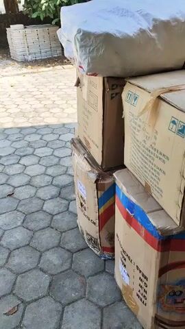 THE VIDEO SHOWS SEVERAL CARDBOARD BOXES STACKED HIGH AND WRAPPED IN PLASTIC, OUTSIDE ON A PAVED PATIO. TO THE LEFT, A PILE OF SQUARE OBJECTS, POSSIBLY TILES OR PAVERS, IS VISIBLE. FURTHER BACK, THERE ARE TREES AND SHRUBS, A BUILDING, AND MORE PAVING. THE SUNNY WEATHER CASTS SHADOWS ON THE GROUND. THE VIDEO IS STILL, WITH NO MOVEMENT OR PEOPLE PRESENT.