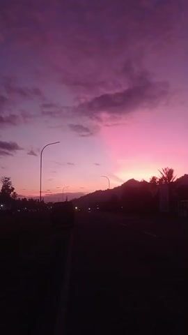 The video shows a dusky sky filled with streaks of purple and pink clouds, possibly a sunset or sunrise. A road stretches into the distance, lined with silhouetted palm trees and a faint mountain range in the background. Streetlights and the headlights of a few vehicles are visible along the road, indicating a low-light environment. The overall scene is calm and serene, with a focus on the atmospheric sky and the quiet road.