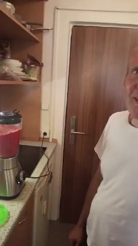 The video shows a kitchen counter with a blender filled with a pink liquid. To the right of the blender, a man in a white t-shirt is visible, looking at the camera and smiling. He appears to be speaking. The kitchen has wooden cabinets above the counter and a wooden door in the background. The blender is on and the liquid is in motion.