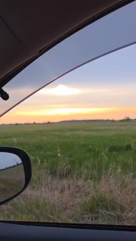 A car is driving along a road through a green field at sunset. The sky is cloudy with a bright orange sun. The side view mirror of the car is visible. Tall grass is near the road.