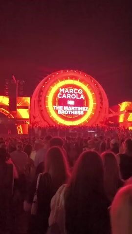 A video shows a large crowd of people at an outdoor nighttime concert. A circular stage structure glows with red and white lights, displaying the text "MARCO CAROLA B2B THE MARTINEZ BROTHERS". Beams of light emanate from the stage into the dark sky. The crowd is densely packed, facing the stage and appears to be enjoying the performance. Some people are raising their hands. The overall atmosphere is that of a lively music festival.