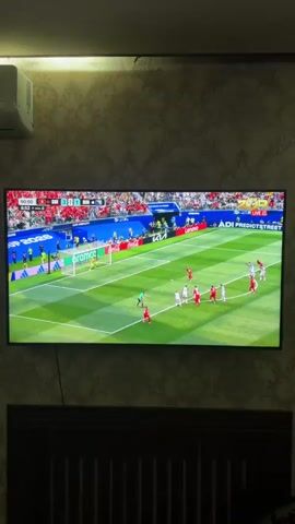 A television screen displays a soccer match. The game is in progress with players in red and white uniforms on a green field with white markings. A scoreboard at the top left shows the time as 90:00 and a score of 0-0. There are spectators in the background. The screen shows a close-up of two players celebrating, one pointing upwards. A graphic on the screen says "GOAL". The television is mounted on a wall with textured wallpaper. Below the television is a dark wooden cabinet. An air conditioner unit is mounted on the wall to the left of the television.