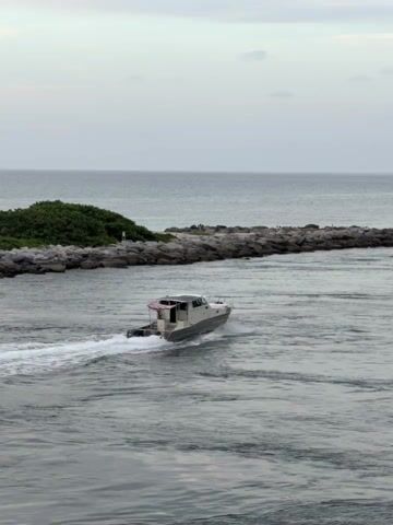 A speedboat travels through choppy water towards a rocky breakwater under a cloudy sky. The boat, grey and white with a small American flag on its stern, leaves a white wake behind it. The breakwater is made of large grey rocks and extends into the ocean, with a small patch of green vegetation on the far left. The water is dark and grey, reflecting the overcast sky. The horizon line between the water and sky is visible in the distance.