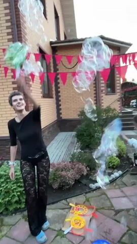 A person with short dark hair and a black shirt is in a backyard, making a lot of bubbles. They are holding a green bubble wand and seem to be having fun. There are red triangular flags strung across the house, and a lot of soap bubbles are floating in the air. There are also some bubble wands on the ground, along with a blue and green bucket. The house is made of brick and has a paved patio in front of it.