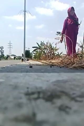 The video begins with a low-angle shot of a gray, textured surface, likely asphalt or concrete, with some dark debris scattered on it. In the background, a woman dressed in a long, magenta hijab and dress stands leaning on a cane. She appears to be elderly. Behind her, there is a pile of dry, straw-like vegetation and several banana trees with lush green leaves. To the left of the woman, a tall, thin pole with what appears to be a spider-like structure at the top is visible. Further in the background, a power line with multiple wires stretches across the sky. The sky is bright blue with some scattered white clouds. The scene suggests an outdoor, possibly rural or semi-rural, environment on a sunny day. The camera angle is very low, emphasizing the foreground surface and creating a sense of looking up at the scene. There are no other people visible in the frame. The woman is stationary, and the camera is also stationary.