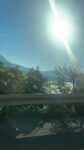 The media file shows a scenic view from a vehicle moving along a road. The sun is bright and high in the sky, indicating it's likely daytime, possibly mid-morning or early afternoon. The weather appears to be clear and sunny.

The scene features a large body of water, likely a bay or lake, with mountains in the background. The water is calm and reflects the bright sunlight. Lush green trees and vegetation line the roadside, with some branches and leaves intruding into the foreground of the shot. A metal guardrail runs along the edge of the road, separating it from the drop-off towards the water. The road itself is paved and has visible tire tracks and shadows cast by the trees.

The perspective suggests the footage is captured from a moving vehicle, looking out to the left. The bright sunlight creates lens flares and a strong glare, obscuring some finer details. The overall emotion conveyed is one of a peaceful and beautiful drive through a natural landscape. There is no visible text within the media file. The location context provided is Provodina, Montenegro, which aligns with the mountainous coastal scenery.