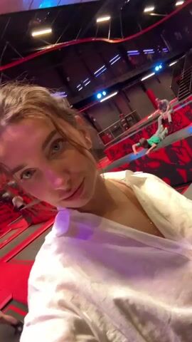 In a brightly lit indoor trampoline park, a woman with brown hair pulled back, wearing a white shirt, is recording a selfie video. The camera is close to her face and she has a slight smile. The background is filled with various trampoline setups, including red and black patterned areas, light fixtures, and other people moving around. There are also neon lights and colorful displays visible in the background. The camera moves slightly as she appears to be walking or turning.