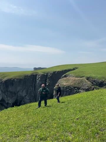 Two men are walking on a grassy field atop a cliff. The man in front is wearing a dark green hooded sweatshirt and jeans. The man behind him is wearing a black shirt and jeans. The man in front stops and looks to his left. The camera pans to the left to reveal a rock arch in the water next to the cliff. The camera continues panning to the left across the water and the island in the distance. The sky is blue with some white clouds.