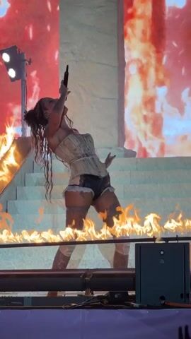 A woman with dark, messy hair tied up performs on a stage, wearing a light-colored corset, black shorts, and thigh-high stockings. Flames are visible around her and on a raised platform behind her. The background features a large screen displaying a fiery image and architectural elements resembling a large column and stairs. A spotlight is visible on the left side of the stage. The woman is holding a microphone and appears to be singing or speaking into it. There are also stage lights and equipment visible in the foreground and on the side.