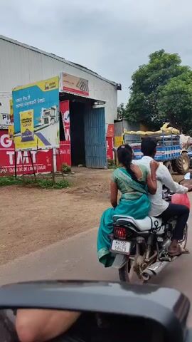 The video captures a scene on a street, likely in India. In the foreground, a motorcycle is being ridden with two people on it. The rider is a man wearing a white shirt. The passenger, a woman, is wearing a green sari and is seated behind the man, with her arm around his shoulder. The motorcycle has a license plate that is partially visible. In the background, there is a building with a red and white facade, which appears to be a commercial establishment, possibly a store or warehouse. Signs on the building are visible, including one with yellow and red lettering that reads "GEL". A blue truck with yellow items stacked in its bed is parked to the right of the building. Another vehicle, possibly a tractor, is also visible further in the background. The ground is wet and has puddles, indicating recent rain or a wet environment. The sky is overcast. The camera appears to be mounted inside a vehicle, as a portion of a side-view mirror is visible in the bottom of the frame. The overall impression is a snapshot of everyday life in a roadside commercial area.