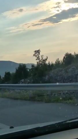 This is a short video clip filmed from a car moving along a road. The view is of a scenic landscape with rolling hills and mountains in the distance. The sun is setting, casting a warm glow across the sky, with scattered clouds. The foreground shows a guardrail and the road surface, indicating travel. The overall mood is peaceful and serene, evoking a sense of journey and natural beauty. The time of day is sunset, and the weather appears fair. The location is identified as Brestnitsa, Bulgaria. There is no visible text in the media.