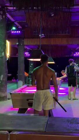 In this video, a man with a tattoo on his back and wearing white shorts stands with his back to the camera, holding a pool cue. He is at a pool table in what appears to be an outdoor bar or entertainment venue. The scene is illuminated by colorful lights, including blue and pink, suggesting it is nighttime or evening. Palm fronds hang from the ceiling, and a thatched roof is visible.

Another man is seen playing pool at a table in the background, bent over to take a shot. Further in the background, a woman is visible near a bar area. The setting seems to be a tropical location, consistent with the mention of Ko Tao Subdistrict, Thailand, with outdoor seating and a relaxed, possibly party atmosphere. The overall mood appears casual and social, with people engaged in leisure activities.