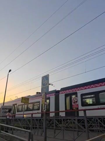The video shows an outdoor scene at dusk with a clear sky, transitioning from light blue overhead to orange and pink at the horizon. Two trains are visible on parallel tracks. A red and white train, possibly a Moscow Central Diameter (MCD) train, is partially visible on the right, stationary or moving slowly. Another white train with red lights on its front approaches from the left, passing by the viewer from left to right. Overhead power lines crisscross the sky. A tall lamppost stands in the mid-ground. To the left, a person is standing on a platform or elevated area near a sign with Cyrillic text, largely silhouetted against the sunset. A blue circular sign with a white 'M' logo is prominently displayed between the tracks. A metal railing is in the foreground.