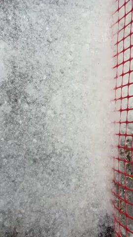 A close-up, vertical video shows white material, possibly hail, falling onto a dark surface, possibly pavement. The material is granular and appears to be accumulating. On the right side of the frame, a red mesh fence is visible. The video is dark, with the primary light source appearing to be the falling white material itself.