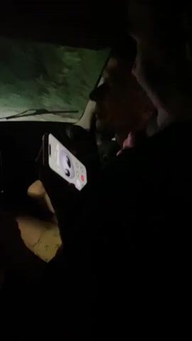 Two individuals are in the dark interior of a vehicle. One person, in the foreground and on the left, holds up a smartphone. The screen of the phone is illuminated, displaying a contact card with a large circular profile picture and text. The other person is partially visible behind and to the right of the first individual, looking towards the phone. The background outside the vehicle is dark, with some indistinct green or grey illuminated areas, possibly foliage or a landscape. The scene suggests a moment of interaction involving the smartphone, possibly a call or checking information.