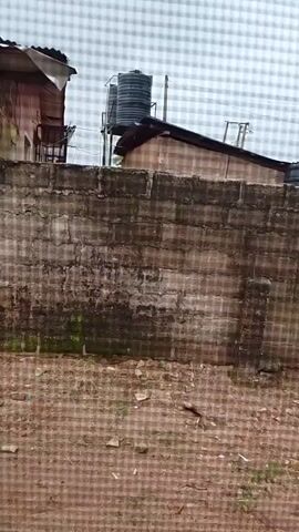 The video shows a rough, textured wall made of concrete blocks, partially covered in green moss. The ground in front of the wall is composed of dirt and debris. Behind the wall, there are buildings with corrugated roofs and water tanks on top. The sky appears overcast. The camera seems to be moving, indicated by the slight blur and shifting perspective.