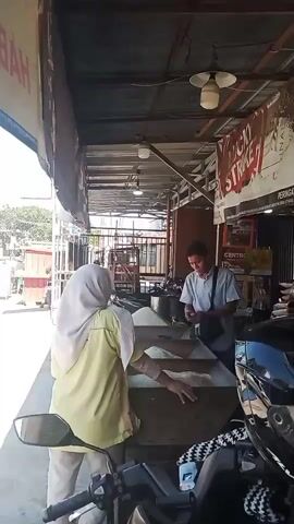 A man in a light blue shirt and dark pants is standing behind a counter, holding a smartphone and a pen. He is looking down at something in his hands. In front of him, a woman in a yellow shirt and a white hijab is reaching into a large wooden bin filled with white rice. The bin is open and the rice is piled high. Behind the man, there is a sign with red lettering that reads "LUCKY STRIKE." The scene appears to be outdoors, possibly at a market stall, with a motorcycle parked in the foreground. The lighting is bright, suggesting daytime.