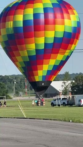 A large, brightly colored hot air balloon with a rainbow pattern is being prepared for flight on a grassy field. The balloon is partially inflated, with its lower section attached to a basket where several people are standing. A person appears to be operating a burner, directing flames into the balloon. In the background, there are trees, a building, a white pickup truck, and a trailer. The sky is clear and blue.