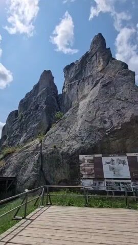 This video shows a scenic view of a rocky cliff formation with a wooden walkway in the foreground. Several people are walking on the walkway, some of them are taking pictures. There is a sign with information near the cliff. The sky is blue with white clouds. The environment appears to be a natural park or tourist attraction.