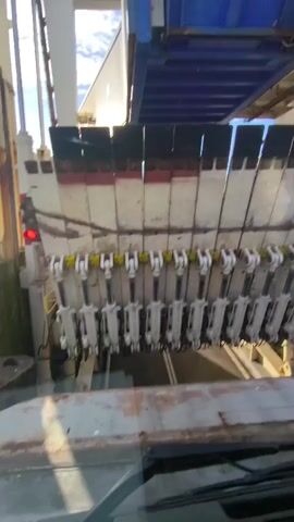 The video shows a close-up view of a ramp mechanism. Numerous hydraulic cylinders are visible, arranged in a horizontal line and attached to a series of metallic panels. These panels are angled upwards and appear to be part of a deployable ramp. The cylinders are white with yellow accents. Above the ramp mechanism, a section of a blue structure and a white building are visible against a bright sky. The overall scene suggests an industrial or maritime setting where a ramp is being operated.