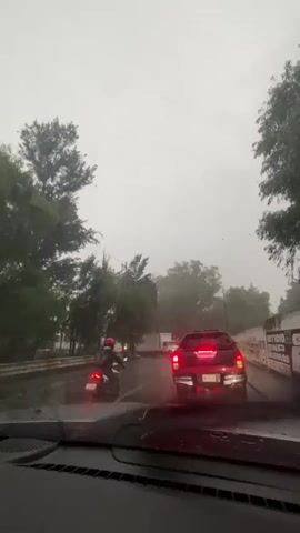 The video captures a road scene from inside a vehicle, looking forward through a rain-splattered windshield. Heavy rain is actively falling, creating wet road conditions and reducing visibility, accompanied by the distinct sound of a windshield wiper. Traffic is present on the multi-lane road, with vehicles either moving slowly or stopped.

In the immediate foreground, the illuminated red tail lights of a black pickup truck are prominent. Its license plate clearly displays "PBV-55-84." Further ahead in the traffic, a white box truck and a motorcycle with a rider wearing a red helmet can be seen.

The road is flanked by green trees on both sides, and on the right, a wall is visible, featuring partially obscured text that includes the word "SIENTE." The sky is overcast due to the storm, suggesting daytime conditions darkened by the heavy weather. The scene is set in Cuautitlán Izcalli, Mexico.