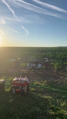 The video shows a large agricultural drone taking off from a field at sunrise or sunset. The drone, which has multiple rotors and a liquid payload container, rises steadily into the air against a blue sky marked with contrails. In the foreground, a red and black generator sits on the ground with a green hose leading towards the drone. The field is green, and the sun is low on the horizon, casting a warm glow.