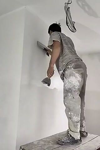 A man is standing on a platform and plastering a wall with a trowel. He is wearing a grey t-shirt and grey pants that are splattered with white paint. He is also wearing grey crocs. There is a white ceiling and a white wall. There is also a wire hanging from the ceiling.