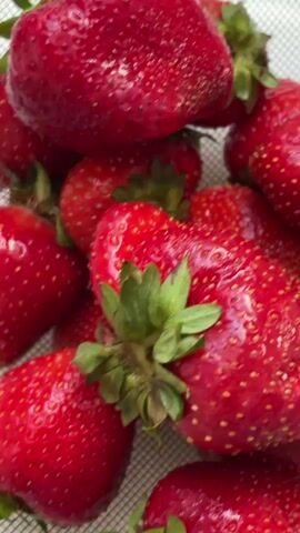 This is a close-up video of a pile of fresh strawberries. The strawberries are bright red with visible yellow seeds and green stems. The camera is positioned directly above the strawberries, and the focus shifts slightly throughout the video, creating a soft, blurred effect. The background appears to be a light-colored, textured surface, possibly a mesh colander or fabric. The lighting is bright, highlighting the glossy texture of the berries. No people or actions are visible, only the static arrangement of the fruit.