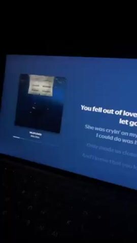 A laptop sits in darkness, its screen illuminated and displaying music lyrics and album art. The lyrics read "She was cryin' on my shoulder. All I could do was hold her. Only made us closer. And I know that you know you don't need to remember. I should put it all behind us." The album art shows what appears to be a human heart against a dark blue, watery background. The title of the song, "WILDFLOWER," and artist, Billie Eilish, are displayed on the screen. The laptop keyboard is visible, but the specific model cannot be determined without further visual cues. The overall atmosphere suggests a focus on the screen content and the musical selection.