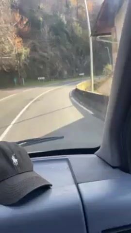 The video shows a view from inside a vehicle driving on a winding road. The road has white lines and directional signs. The environment outside consists of trees and vegetation along the roadside. The interior of the vehicle is visible, including the dashboard and a baseball cap with a logo. Sunlight shines through the windshield.