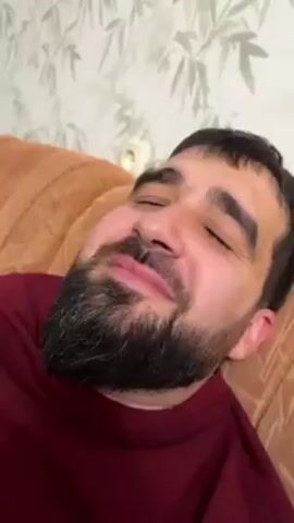 Here's a plain text description of the video:

A man with a beard is lying on a couch. He is wearing a maroon shirt. His eyes are closed and he has a slight smile on his face. Someone is using blue plastic scissors to gently trim his beard. The background shows a wall with a light floral pattern and an electrical outlet. The couch is light brown. Music is playing in the background.