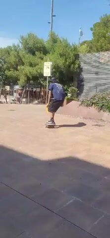 A person on a skateboard is seen performing a trick near a yellow curb. The skateboarder, dressed in dark clothing, is captured mid-air as they jump over the curb. The surrounding environment appears to be an outdoor public space with paved ground, trees, bushes, and a fence in the background. The sky is clear and blue, indicating a sunny day.