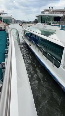 This media file captures a dynamic scene of two passenger boats docked side-by-side on a cloudy day in Budapest, Hungary. The foreground is dominated by the railing of one boat, giving a close-up view of its structure. Beyond this, the second boat, labeled "GONDOLA," is prominently featured. This gondola-style boat has a sleek white exterior with large, tinted glass windows that reveal rows of blue seats inside, indicating it's ready to transport passengers.

Two individuals are visible on the gondola boat: one is standing near the entrance, possibly attending to boarding or disembarking passengers, while another person is seated inside. The water between the two boats is choppy, suggesting movement or a slight swell. In the background, a bridge and various buildings are discernible, further confirming the Budapest setting. The sky is overcast, hinting at a possibly warm but not sunny day. The overall impression is one of an active port or docking area, with the boats poised for their next journey. The visible text on the boat reads "GONDOLA" and what appears to be "DUNA," likely referring to the Danube River.