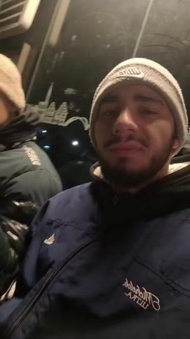 Two young men are riding on what appears to be public transportation, possibly a bus or train, at night. Both are wearing beanies and winter jackets. The man in the foreground has a short beard and is wearing a navy blue jacket with white writing. The man behind him has his fist clenched and is wearing a dark jacket. The video appears to be taken by the man in the foreground, facing the camera. Outside the window, lights and indistinct urban elements are visible. The lighting is somewhat dim and uneven.
