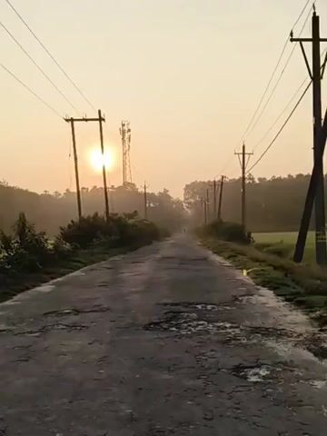 The video shows a rural road at sunrise. The sun is low in the sky, casting a warm glow and creating a hazy atmosphere. The road is paved but appears to be in poor condition, with numerous potholes. On either side of the road, there are utility poles with power lines and a communication tower in the distance. Dense vegetation lines the sides of the road, and trees form a backdrop. The overall impression is one of a quiet, early morning scene in a rural setting.