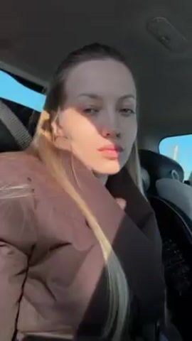 A woman is recording a video of herself inside of a vehicle. She has blonde hair and is wearing a brown coat. The sun is shining through the window, creating shadows on her face. She moves slightly in the seat to adjust her coat. A car seat is visible behind her.