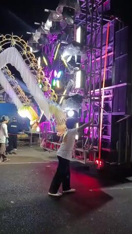 At night, a person in a white t-shirt and black pants stands in front of a large sound system with many lights. The person is facing away from the camera and appears to be interacting with the lights, possibly dancing or operating them. The sound system is elaborate, with multiple speakers stacked on top of each other and a complex structure of metal beams and lights. There are decorative elements, possibly part of a stage or a float, to the left of the sound system, including white fabric draped in an arch. Some other people are visible in the background, standing near tables. The ground is dark, and the lighting from the sound system illuminates the scene with various colors, including white, pink, and purple.