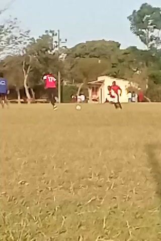 The video shows a soccer game on a grassy field. Several players wearing different colored jerseys, including red, blue, and yellow, are actively playing, running and kicking the ball. The goalposts are visible at the end of the field. Trees surround the field. A small building can be seen in the background on the right. The video is somewhat blurry.