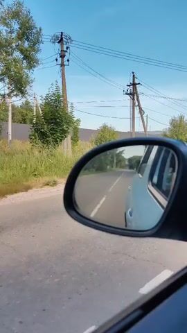 The video shows a view from inside a car, looking out the side mirror. The mirror reflects a road with a white line down the middle, and a light-colored vehicle traveling ahead. The scenery outside the car, visible to the left of the mirror, includes a grassy roadside, trees, and a power pole. In the distance, a building with a dark roof and solar panels is visible, along with a small blue structure that appears to be a bus stop. The sky is clear and blue, suggesting it is daytime and the weather is good. The video appears to be taken from a moving vehicle, likely in a rural or suburban area. No people are visible in the frame. The overall mood is calm and observational, capturing a moment of travel.